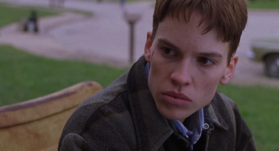 White trans man with short brown hair and paid jacket sits on a beach. He has a semi-healed cut on